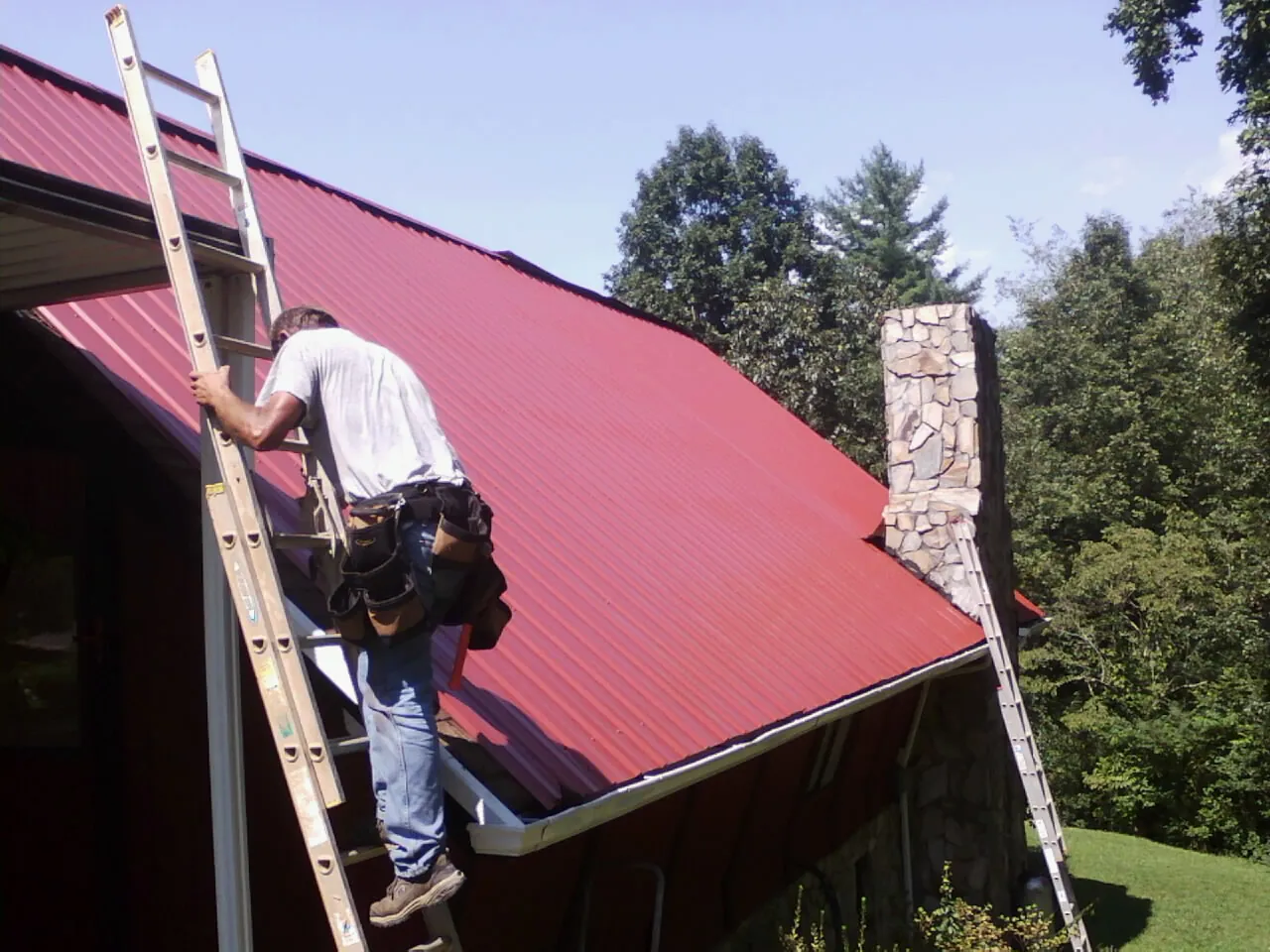 Roofer on a ladder beside a red metal roof and stone chimney — Soffit & Fascia Repair by Roofing Brothers in Stafford Springs